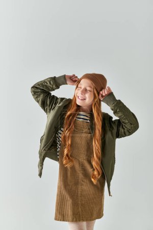 smiling girl with long hair wearing winter hat and standing in dress with jacket on grey background