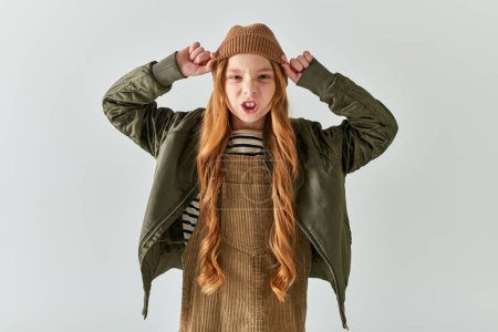 emotional girl with long hair wearing winter hat and standing in dress with jacket on grey backdrop