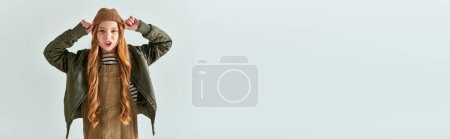 emotional girl with long hair in dress with warm jacket wearing winter hat on grey backdrop, banner