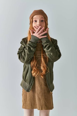 winter fashion, shocked girl with long hair and knitted hat standing in dress and jacket on grey
