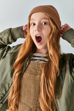 moda de invierno, chica emocional con la boca abierta con sombrero y mirando a la cámara en el fondo gris