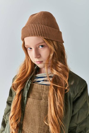 winter fashion, serious girl in knitted hat and outerwear looking at camera on grey backdrop