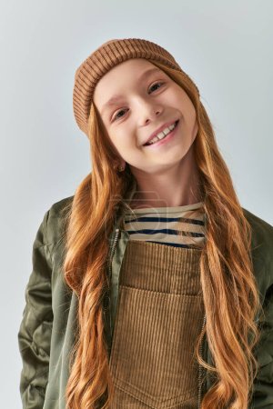 winter fashion, cheerful girl in knitted hat and outerwear looking at camera on grey backdrop