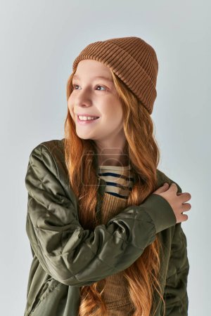 winter fashion, dreamy girl in knitted hat and outerwear looking away on grey backdrop