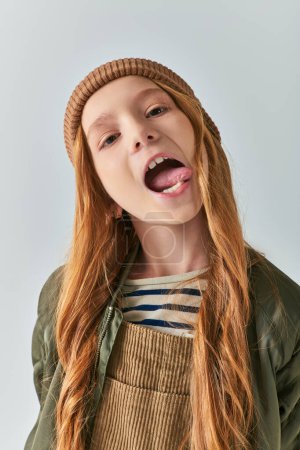 preteen girl in knitted hat and outerwear sticking out tongue and looking at camera on grey backdrop