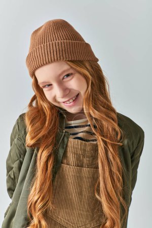 niño con estilo, chica alegre en sombrero de punto y ropa de abrigo mirando a la cámara sobre fondo gris