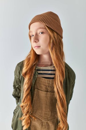 doubtful preteen girl  with long hair posing in winter outfit and knitted hat on grey backdrop