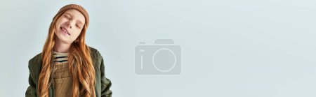 cute girl in knitted hat grimacing and looking at camera on grey backdrop, winter fashion banner