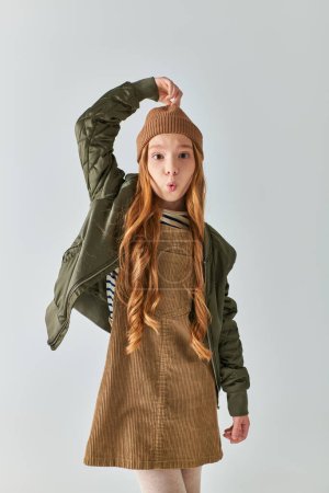 Preteen Girl in stylischem Winteroutfit mit Strickmütze und schmollenden Lippen vor grauem Hintergrund