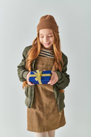 fröhliches Mädchen in stylischem Winteroutfit mit Hut, der verpacktes Weihnachtsgeschenk auf grauem Hintergrund hält