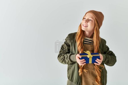 positives Mädchen im stylischen Winteroutfit mit Hut, der verpacktes Weihnachtsgeschenk auf grauem Hintergrund hält