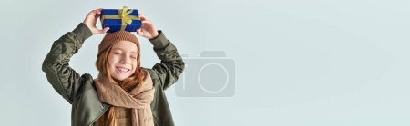 Photo for Smiling girl in stylish winter outfit with hat holding Christmas present above head on grey, banner - Royalty Free Image