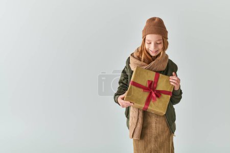 cheerful girl in trendy winter outfit with knitted hat holding Christmas present on grey backdrop