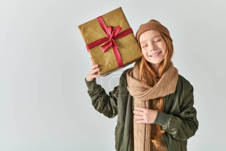 joyous preteen girl in winter outfit with knitted hat holding Christmas present on grey backdrop