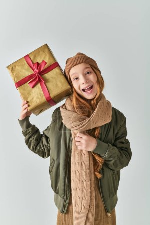 excited preteen girl in winter outfit with knitted hat holding Christmas present on grey backdrop