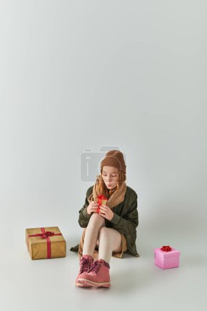 niedliche preteen Mädchen im Winter-Outfit mit Strickmütze hält Weihnachtsgeschenk und sitzt auf grau