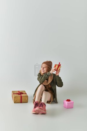 happy preteen girl in winter outfit with knitted hat holding Christmas present and sitting on grey