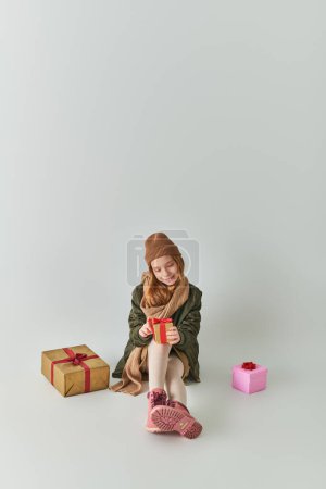 fröhliches Mädchen im Winteroutfit mit Strickmütze, das ein Weihnachtsgeschenk in der Hand hält und auf grau sitzt