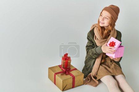 smiling preteen kid in stylish outfit with winter hat holding Christmas present and sitting on grey