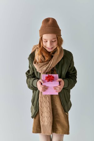 Photo for Happy preteen kid in stylish outfit with winter hat holding Christmas present and standing on grey - Royalty Free Image