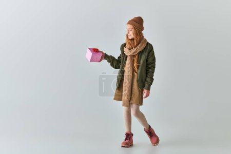 alegre niña preadolescente en ropa de invierno y sombrero celebración de regalo de Navidad y de pie en gris