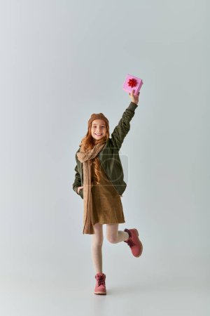 joyous preteen girl in winter clothes and hat holding Christmas present and standing on grey Poster 680988956
