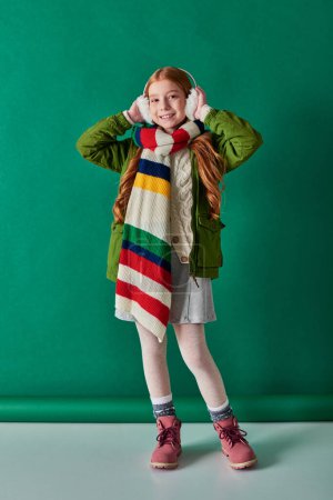 Téléchargez les photos : Couches confortables, fille gaie dans des cache-oreilles et foulard rayé debout en tenue d'hiver sur turquoise - en image libre de droit