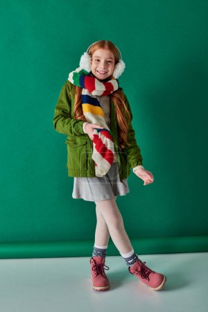 Foto de Chica feliz en orejeras y bufanda de pie en traje de invierno sobre fondo turquesa, capas acogedoras - Imagen libre de derechos