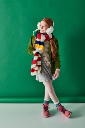 cheerful kid in ear muffs and scarf standing in winter outfit on turquoise backdrop, cozy layers