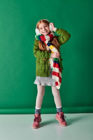 positive child in ear muffs and scarf standing in winter outfit on turquoise backdrop, cozy layers
