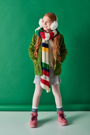 full length, cheerful girl in ear muffs, striped scarf and winter outfit on turquoise backdrop