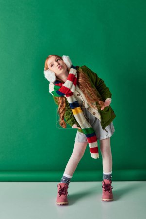 Photo for Full length, preteen girl in ear muffs, striped scarf and winter outfit pouting lips on turquoise - Royalty Free Image