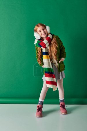 full length, joyful girl in ear muffs, striped scarf and winter outfit on turquoise backdrop