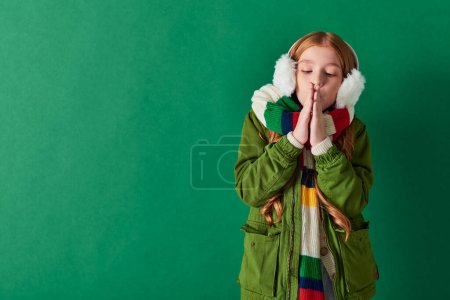 preteen girl in ear muffs, striped scarf and winter outfit warming up hands on turquoise backdrop