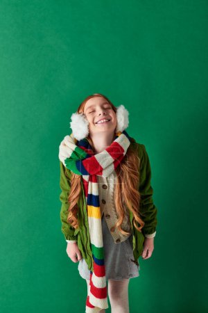 happy preteen girl in ear muffs, striped scarf and winter outfit standing on turquoise backdrop