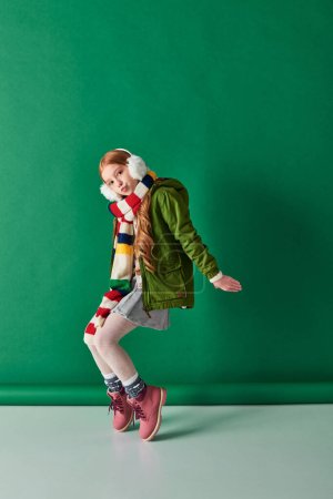 stylisches Preteen Girl in Ohrenschützern, gestreiftem Schal und Winteroutfit posiert vor türkisfarbenem Hintergrund
