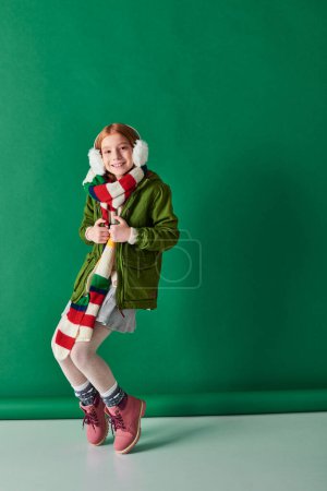 cheerful preteen girl in ear muffs, striped scarf and winter outfit posing on turquoise backdrop
