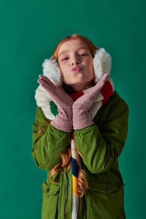 preteen girl in ear muffs, striped scarf and winter outfit pouting lips on turquoise, air kiss