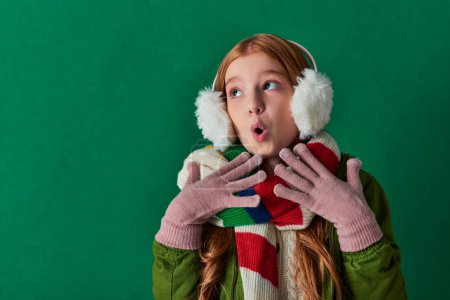 shocked preteen girl in ear muffs, striped scarf and winter outfit gesturing on turquoise backdrop