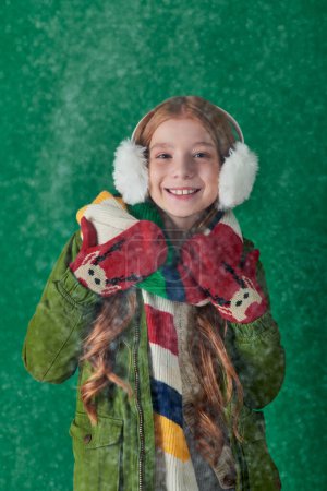 cheerful girl in ear muffs, striped scarf and winter attire standing under falling snow on turquoise