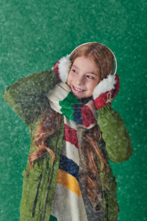 happy girl in ear muffs, striped scarf and winter attire standing under falling snow on turquoise