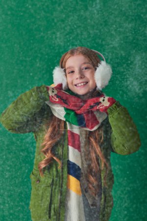 positive kid in ear muffs, striped scarf and winter attire standing under falling snow on turquoise