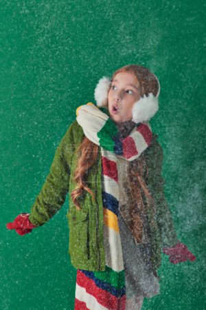 shocked girl in ear muffs, striped scarf and winter attire standing under falling snow on turquoise