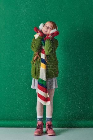 fröhliches Mädchen in Ohrenschützern, warmem Schal und Winterkleidung, das unter fallendem Schnee auf Türkis steht