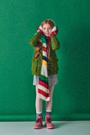 fröhliches Mädchen in Ohrenschützern, warmem Schal und Winterkleidung, das unter fallendem Schnee auf Türkis steht