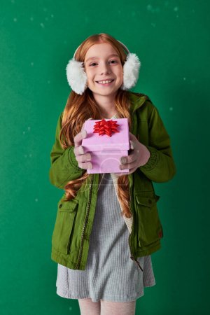 fröhliches Mädchen in Ohrenschützer, Schal und Winterkleidung mit Weihnachtsgeschenk unter fallendem Schnee