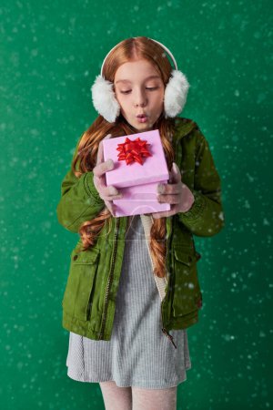 niña preadolescente en orejeras, bufanda y atuendo de invierno soplando nieve de regalo de Navidad en turquesa