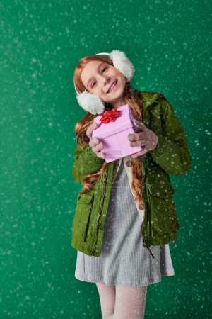 pleased girl in ear muffs, scarf and winter attire holding Christmas present under falling snow