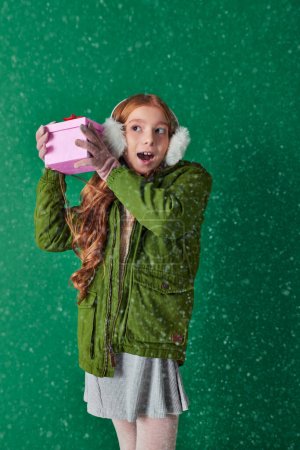 amazed girl in ear muffs, scarf and winter attire holding Christmas present under falling snow