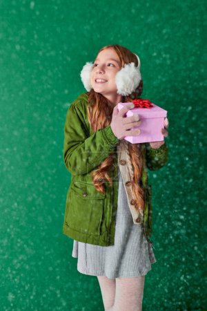 Jahreszeit der Freude, Mädchen in Ohrenschützern und Winterkleidung mit Weihnachtsgeschenk unter fallendem Schnee